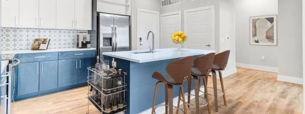 Modern kitchen with blue lower cabinets, white upper cabinets, and a central island with three wooden stools. A fruit bowl sits on the counter. Hardwood flooring and contemporary decor are visible.