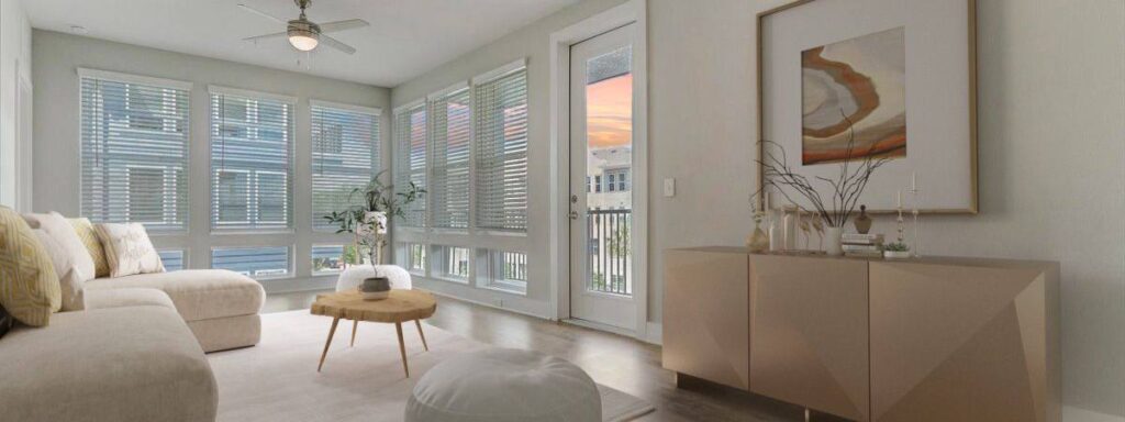 A modern living room with a large beige sectional sofa, a round wooden coffee table, and a tan sideboard. Large windows and a glass door overlook a sunset view.