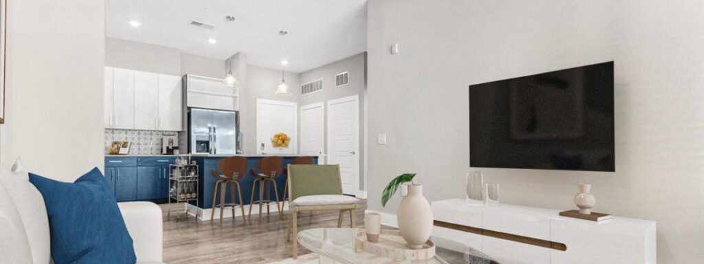 Modern living room with a wall-mounted TV, white sofa with blue pillows, and a view into a kitchen with blue cabinets and bar stools. Light walls and wood flooring create a bright space.