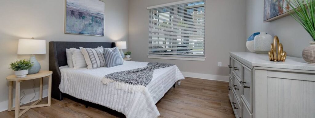 Bedroom with a neatly made bed, gray headboard, two side tables with lamps, a chest of drawers, and a window with blinds. Walls are light-colored, and there are decorative items and art.