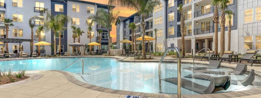 Modern apartment complex with illuminated windows, a swimming pool, lounge chairs, palm trees, and yellow umbrellas at sunset.