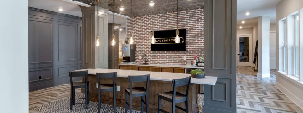 Modern office lobby with a brick accent wall, mounted TV, and a long counter with four chairs. Contemporary lighting and patterned flooring add stylish elements.