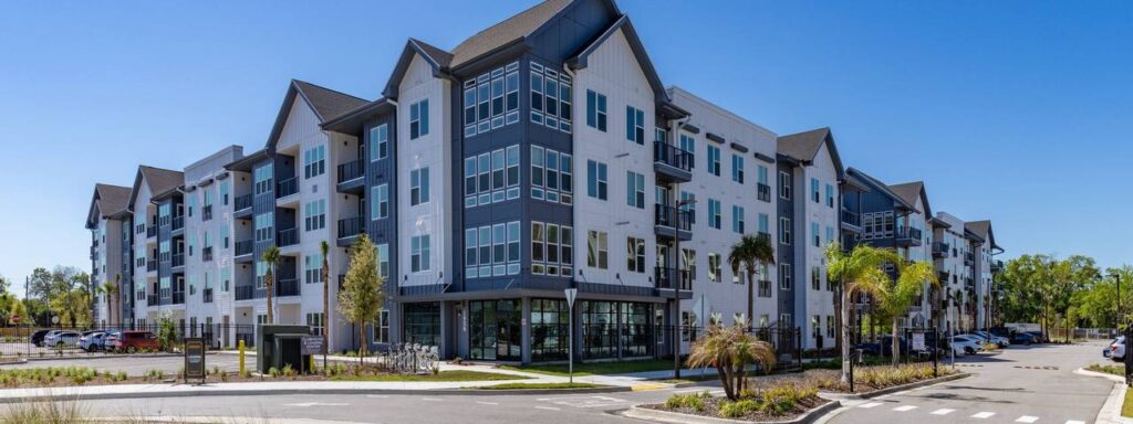 Four-story modern apartment building with glass balconies and a parking area, set against a clear blue sky and surrounded by landscaped greenery.