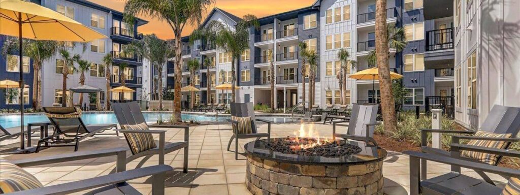 Modern apartment complex with a pool, palm trees, and outdoor seating around a fire pit under a sunset sky.