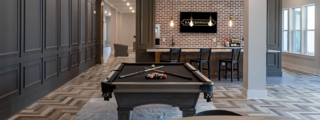 A modern recreation room with a pool table, arranged balls, and cue sticks. Bar area with chairs and a brick wall featuring a TV screen in the background.