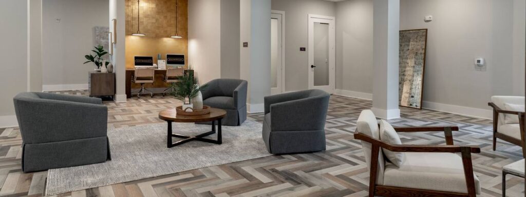 A modern office lobby features gray armchairs, a round wooden table on a rug, and a herringbone patterned floor. In the background, a desk with two chairs is illuminated by overhead lights.