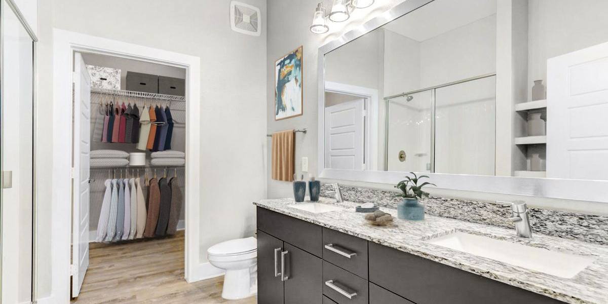 Modern bathroom with a large mirror, granite countertop, dual sinks, and a closet with organized clothes.