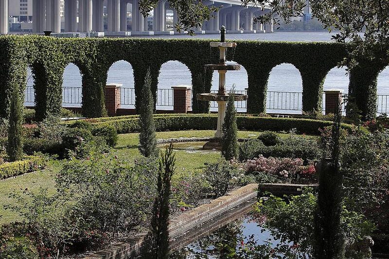 A garden with a central fountain, surrounded by hedges with arches, near a body of water and bridge in the background.