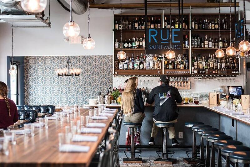 Two people sit at a bar counter in a modern restaurant with hanging lights and shelves stocked with bottles.