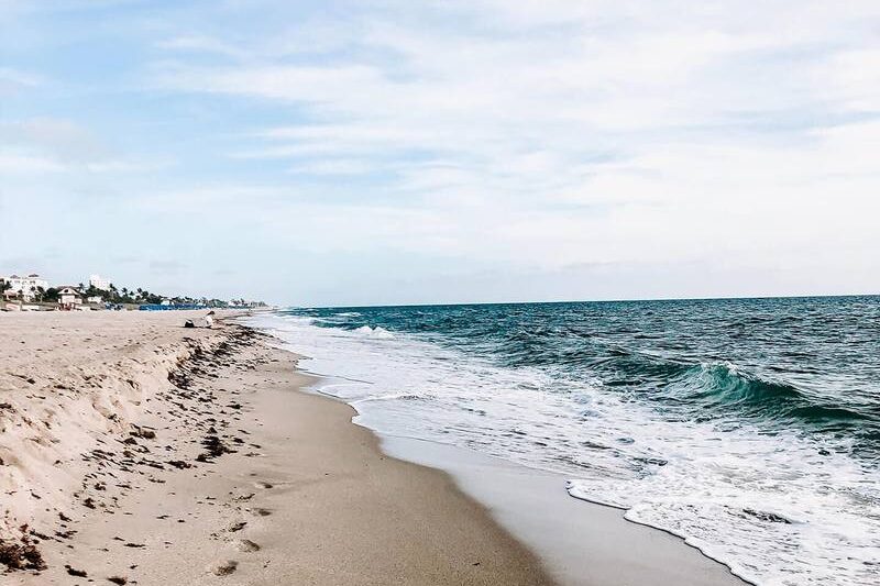 Sandy beach with waves along the shoreline under a partly cloudy sky. Houses and vegetation are visible in the distance.